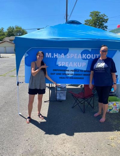 Two women standing under a blue canopy with a mental health awareness banner at an outdoor event.