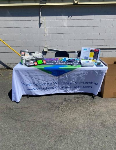 Outdoor table with West Spokane Wellness Partnership banner, displaying brochures, supplies, and informational materials.