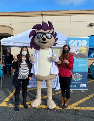 Two women posing with a doctor mascot at an outdoor wellness booth with opioid awareness signage.