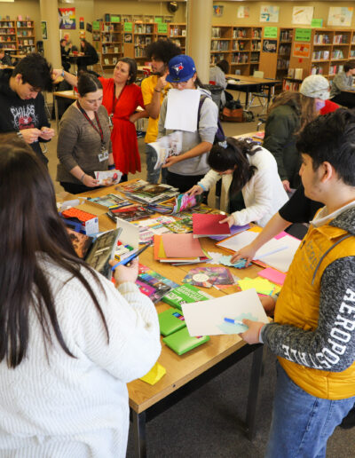 Students gathered around a table in a library working with art supplies and materials during a group activity.