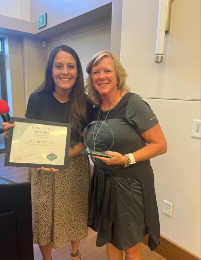 Two women smiling and holding a coalition member certificate and glass award indoors.
