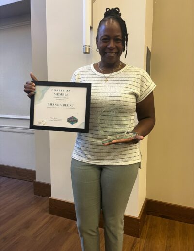 Woman holding a coalition member recognition certificate and glass award while smiling indoors.