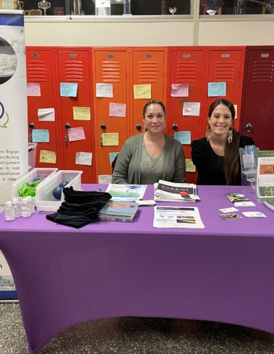 Two women seated behind a prevention outreach table with educational materials and an Operation Engage banner in a school setting.