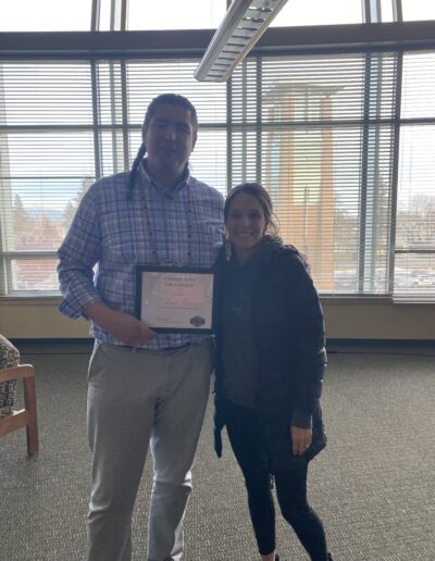 Two people standing indoors, one holding a community champion certificate.