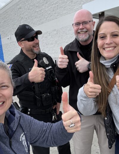 Group of four people smiling and giving thumbs up outside a store, including a police officer.