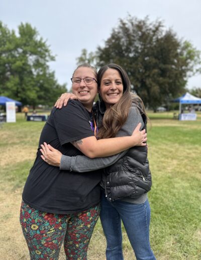 Two women smiling and hugging outdoors at a community event, with tents and trees in the background.
