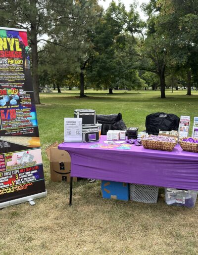 aOutdoor park outreach booth with fentanyl awareness banner, purple table, brochures, and giveaway items displayed.