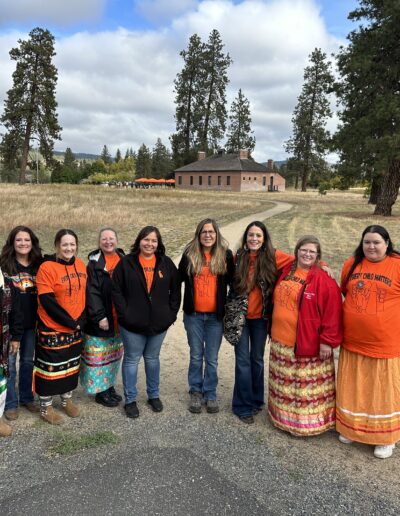 Group of women in orange shirts standing together on a park path with trees and a building in the background.