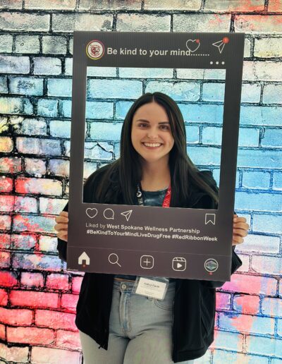 Woman smiling and holding a social media-style frame reading “Be kind to your mind” in front of a colorful brick wall.