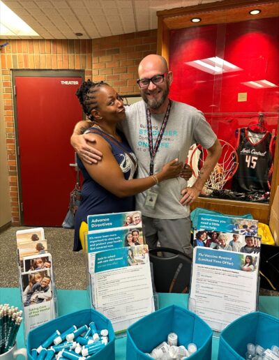 Two people smiling and embracing at a community health booth with informational materials and giveaways in front.