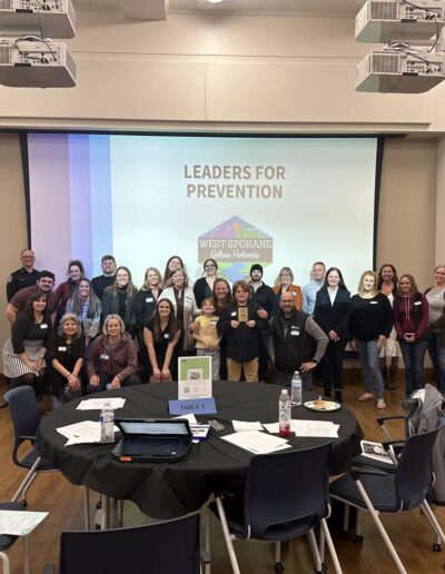 Group photo of West Spokane Wellness Partnership leaders standing in front of a “Leaders for Prevention” presentation screen in a conference room.