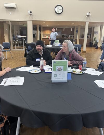 Three people seated at a round table during a community meeting, with others socializing in the background.