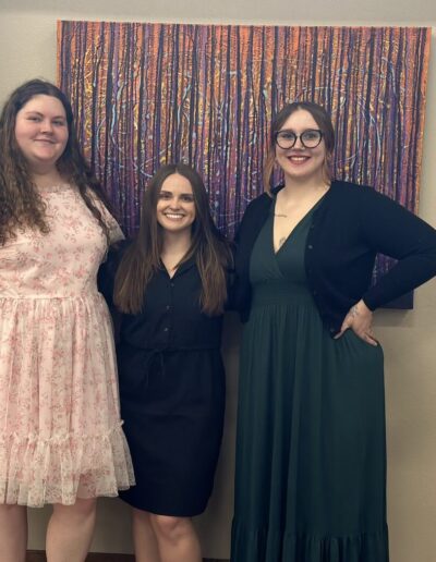Three women smiling and standing together in front of a colorful abstract painting indoors.